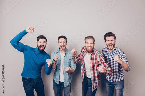 Champions! Four amazed young men are standing and gesturing for the victory on pure background in casual outfit and jeans. They are fans of sports games as football, basketball, hockey, baseball photo