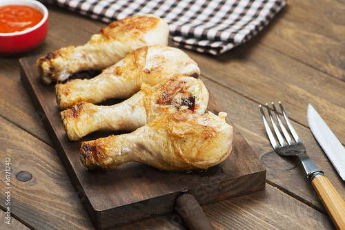 Roasted chicken on wooden board next to bowl with sauce, napkin, fork and knife.