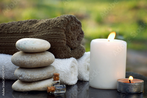 Spa still life with towels, a burning candle, bath oil and massage stones against the backdrop of a green garden in summer