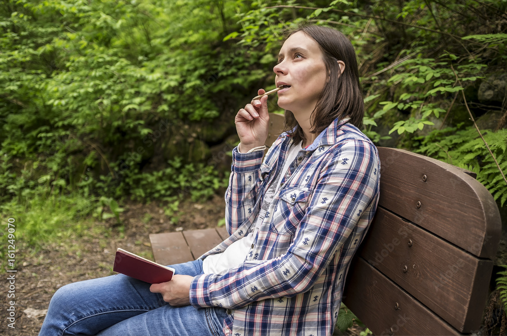 Beautiful woman sitting on a wooden bench in the forest park at the trail and dreamily looks up at the sky, carrying a notebook and a pencil in his mouth. Beautiful woman sitting on a wooden bench in the forest park at the trail and dreamily looks up at the sky, carrying a notebook and a pencil in his mouth.