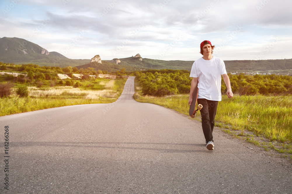 A man in a cap and with a long board in his hands is walking on a country road