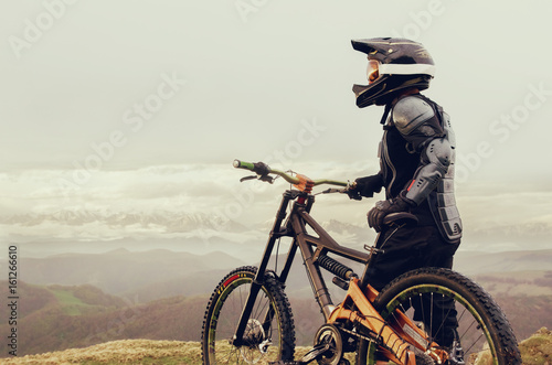 The rider in the full-face helmet and full protective equipment on the mtb bike stands on a rock against the background of a ridge and low clouds