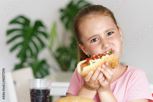 Young cute european girl in t-shirt is eating unhealthy food like hot dog and chips
