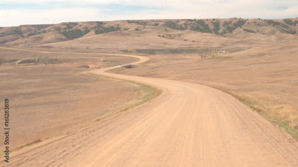 FPV: Driving along the empty dirt road through dry African like grassland. Traveling across the grassy desert during drought on a hot sunny day. Road trip along the winding road across United States. FPV: Driving along the empty dirt road through dry African like grassland. Traveling across the grassy desert during drought on a hot sunny day. Road trip along the winding road across United States.