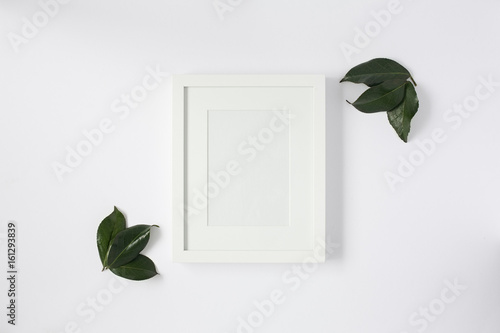 Mock up of white photo frame with leaves on a white desktop. Flat lay.