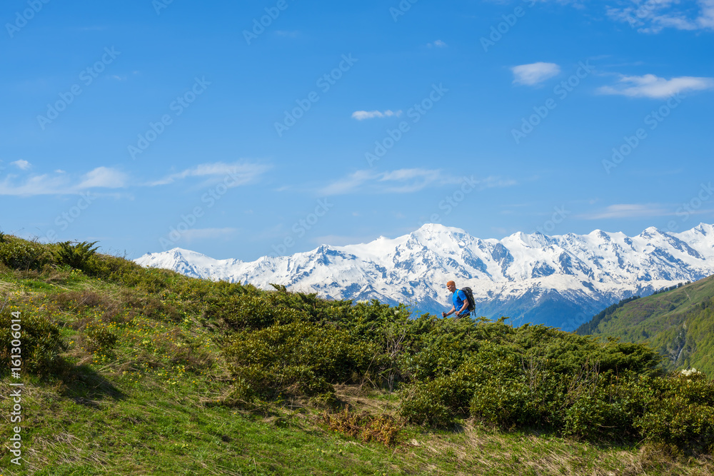 Traveler ascends the green slope against the background of snow-capped mountain peaks