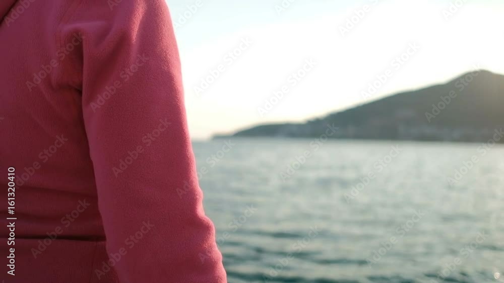 woman sitting on deck by water, looking into distance. Shooting from back attractive female in pink sweater, black pants on bank of lake on stone pier, looking on sunset. beautiful lady with brown woman sitting on deck by water, looking into distance. Shooting from back attractive female in pink sweater, black pants on bank of lake on stone pier, looking on sunset. beautiful lady with brown