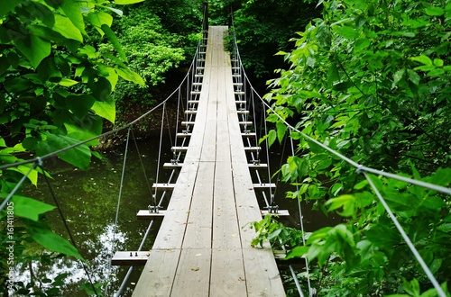 Small wood pedestrian suspension bridge with steel cables over a river in the woods © eqroy