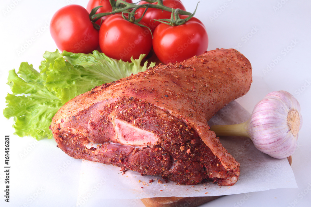 Fototapeta premium Raw pork knuckle and vegetables, garlic, tomatoes spices and lettuce leaves on a cutting board. Selective focus. Ready for cooking.