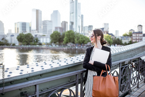 Portrait of a young businesswoman with laptop standing on the bridge in Frankfurt city