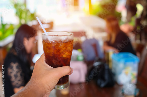 Close up, Hand holding glass of cola with ice cubes on blurred image of working women are planning and meeting at park. Copy space and texture.