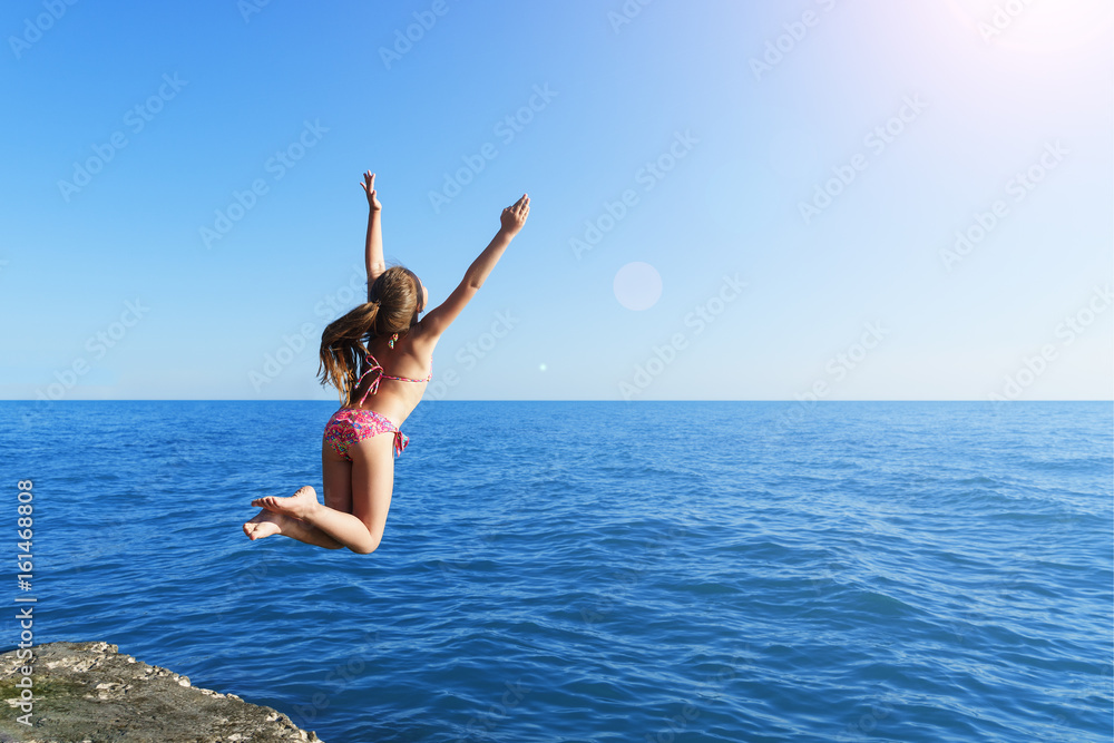 young european cute girl is jumping and flying to the calm blue sea from concrete breakwater towards summer soft sun