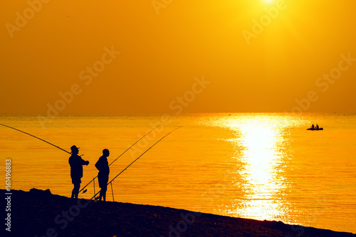 Silhouettes of fishermen with fishing rods on the beach. A boat with fishermen in the sea. Bright colorful sunset photo, dawn. Beautiful marine species. 