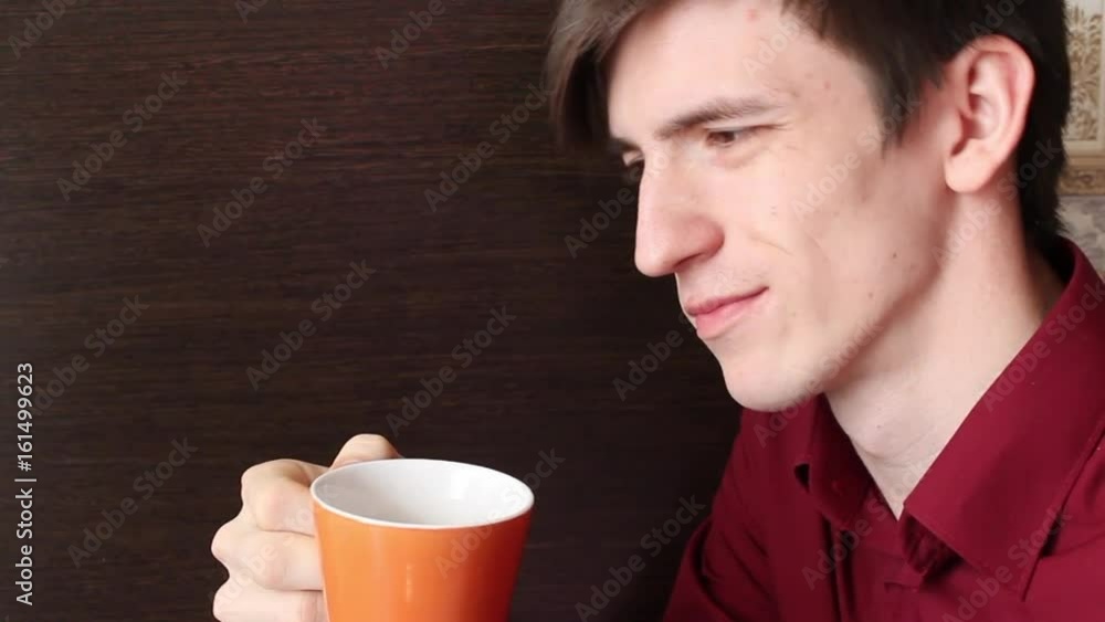 A young guy smiles, takes a slow sip from an orange mug A young guy smiles, takes a slow sip from an orange mug