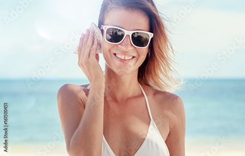 Close up side portrait of a beautiful young woman on holiday on a beach holding a sea shell to her ear and smiling, listening to the sound of the ocean against blue sky. Outdoors lifestyle