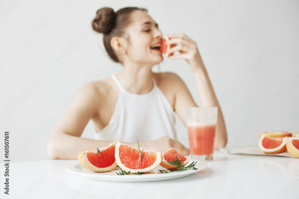 Cheerful young girl smiling sitting at table eating piece of grapefruit over white wall. Healthy food concept. Cheerful young girl smiling sitting at table eating piece of grapefruit over white wall. Healthy food concept.