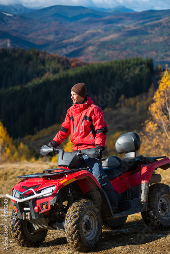 Male rider in jacket and hat on a red ATV on mountain roads on a sunny day. Beautiful landscape of forest, mountains and blue sky