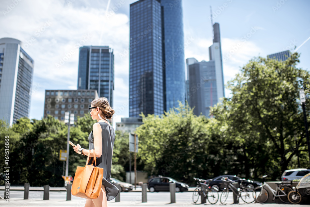 Lifestyle portrait of a businesswoman walking with phone and bag in the modern district in Frankfurt city