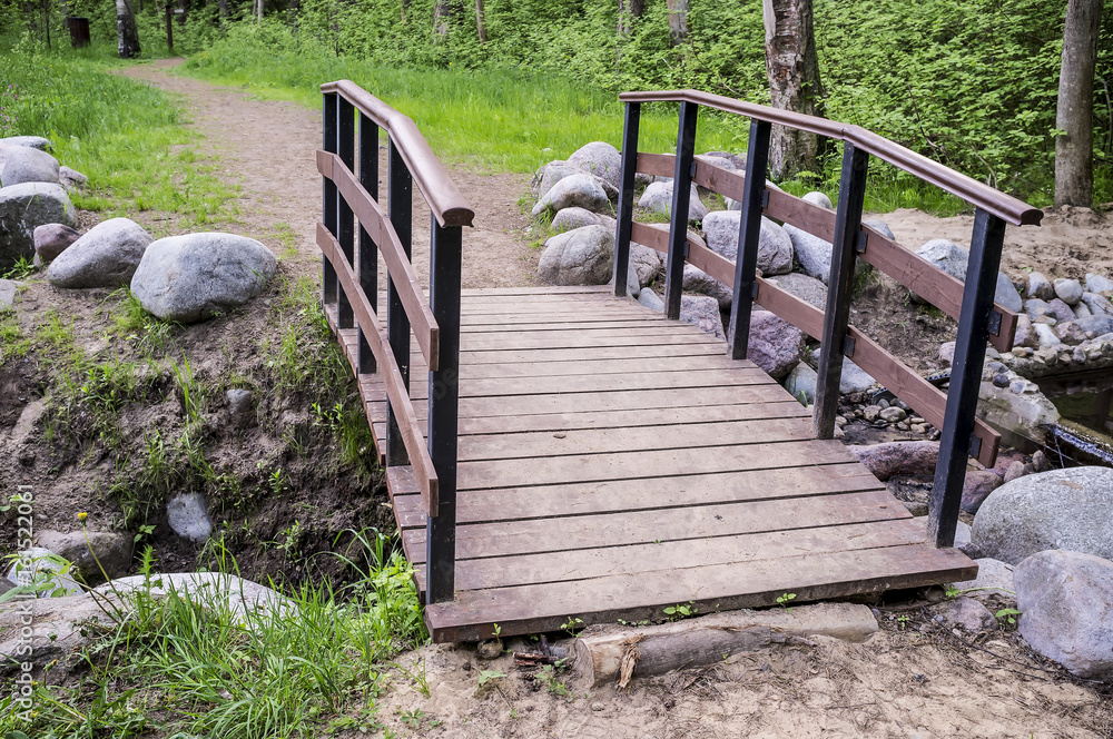 Trail leads to a small wooden bridge over the creek shore is lined with stones, in the summer forest. Trail leads to a small wooden bridge over the creek shore is lined with stones, in the summer forest.