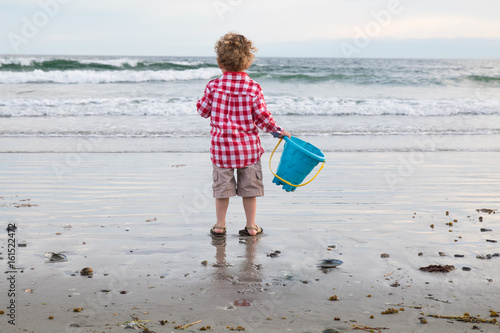A young boy with curly blonde hair is at the beach with his pail. The little child is holding his favorite pail, ready to play at the beach in the afternoon. He is happy. Vacation, summer break