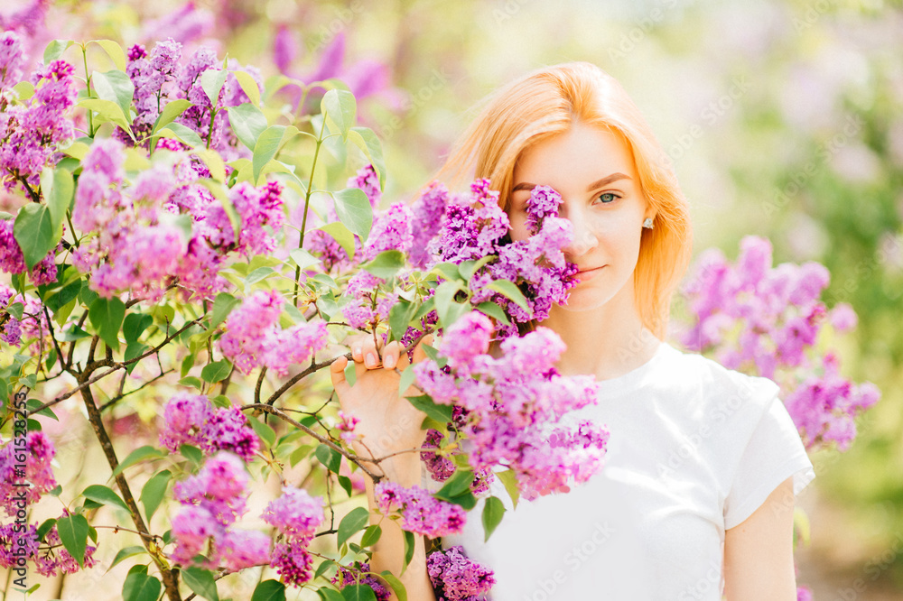 Portrait of young attractive cute redhair girl outdoor in park in blooming lilac bushes in summer sunny day. Resting among paradise flowers. Fragrance and fresh smell. Tenderness, innocence, airness. Portrait of young attractive cute redhair girl outdoor in park in blooming lilac bushes in summer sunny day. Resting among paradise flowers. Fragrance and fresh smell. Tenderness, innocence, airness.