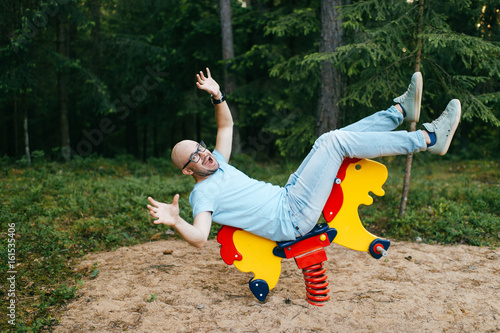 Canvas-taulu Adult bald man in trendy clothing with expressive emotions riding metallic bungee rocking-horse on playground, playing the fool, making scary funny faces in warm weather