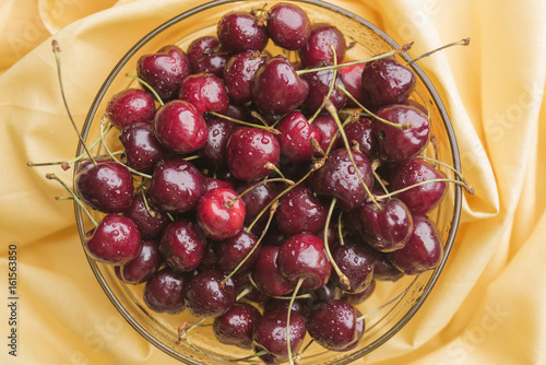 Ripe bright cherry with droplets of water in a transparent bowl on a yellow tablecloth photo