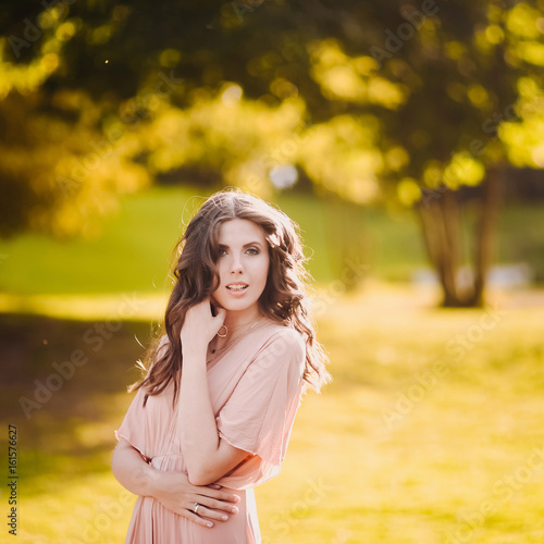 Portrait of a beautiful attractive lady of a young slender brunette girl with long dress walking through a green park with flowers at sunset lifestyle