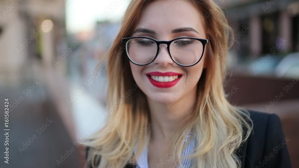 Extreme close up view of a pretty blonde woman in a stylish look, with red lipstick and glasses standing in the city center and charmingly smiling towards the camera. Natural beauty, being happy. Extreme close up view of a pretty blonde woman in a stylish look, with red lipstick and glasses standing in the city center and charmingly smiling towards the camera. Natural beauty, being happy.