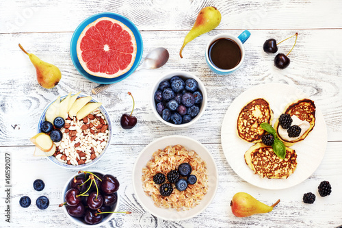Oatmeal and puffed rice cereal with fresh berries, pancakes and grapefruit, cup of coffee and pears on white table. Top view of healthy breakfast. 