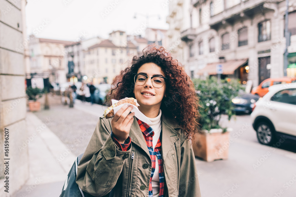Fototapeta premium Young beautiful mixed race woman outdoor in the city eating cannolo - sweet, dessert, pastry concept