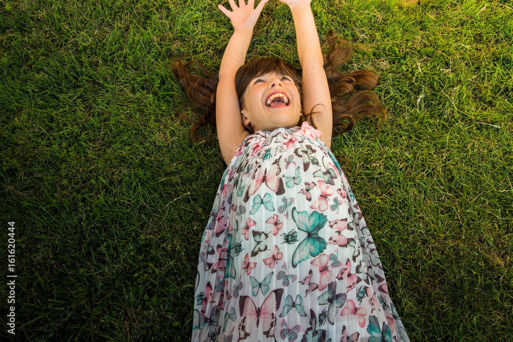 Cheerful little girl lying on green lawn / Joyful little girl enjoying and laughing with hands above her head while laying on the green lawn Cheerful little girl lying on green lawn / Joyful little girl enjoying and laughing with hands above her head while laying on the green lawn