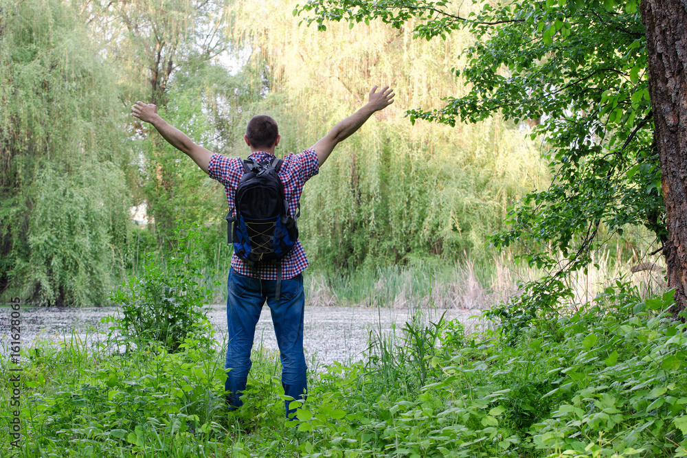 Guy with a backpack is standing on the shore of a forest lake with arms outstretched, a back view