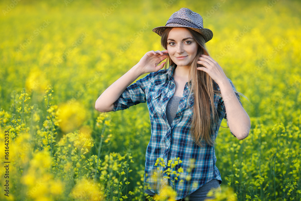 Attractive young blonde woman in blue plaid shirt straw hat enjoying her time on bright colorful blossoming yellow green meadow hard noon sun. Attractive young blonde woman in blue plaid shirt straw hat enjoying her time on bright colorful blossoming yellow green meadow hard noon sun.