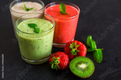 Close-up three glasses with smoothies, banana, kiwi, strawberries, on a black stone surface, decorated with mint, next to lie fresh berries and fruits, a tasty bright dessert, healthy food, restaurant