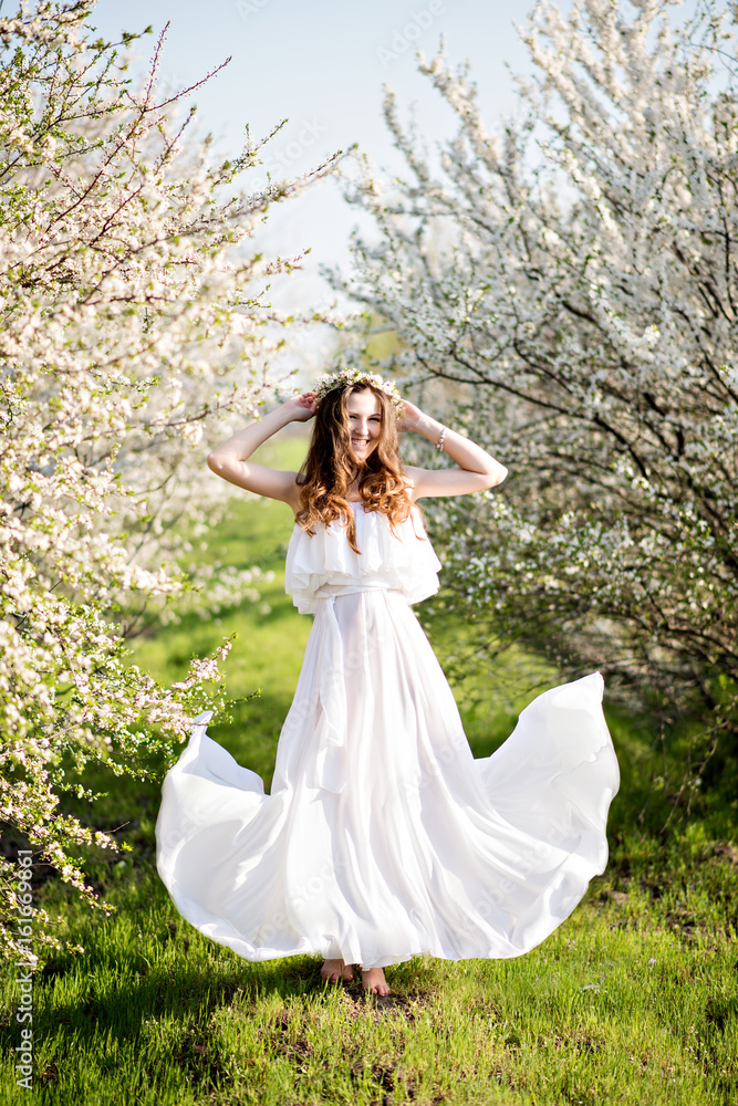 Beautiful young girl in white dress and wreath of flowers on head in blooming gardens