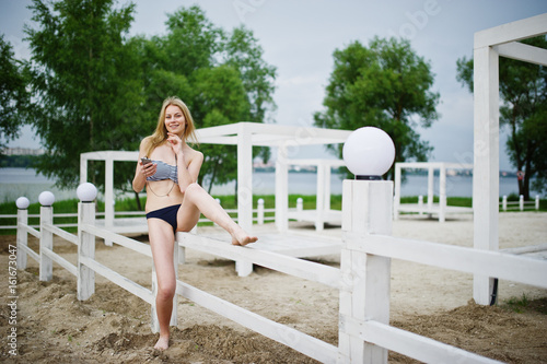 Portrait of a stunning young female model in bikini posing next to the white wooden fence in the park.