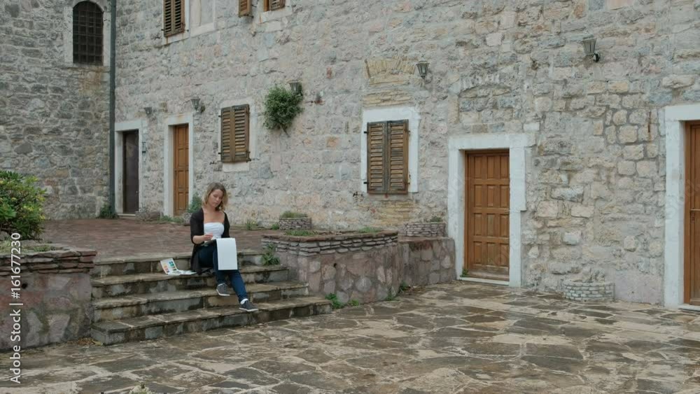 Female artist paints on a white canvas cityscape on the steps. Beautiful lady sitting on the stairs among the stone walls of a European city and makes the brush thumbnail. She looks at an object that Female artist paints on a white canvas cityscape on the steps. Beautiful lady sitting on the stairs among the stone walls of a European city and makes the brush thumbnail. She looks at an object that