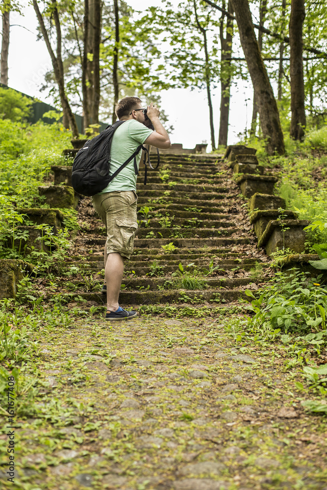 Tourist with a backpack makes summer forest pictures in the park on the retro camera, walking through the old cobbled path and stairs in the moss.