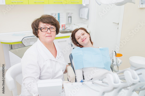 Portrait of middle aged female dentist with smiling patient sitting at dental chair  both looking at camera