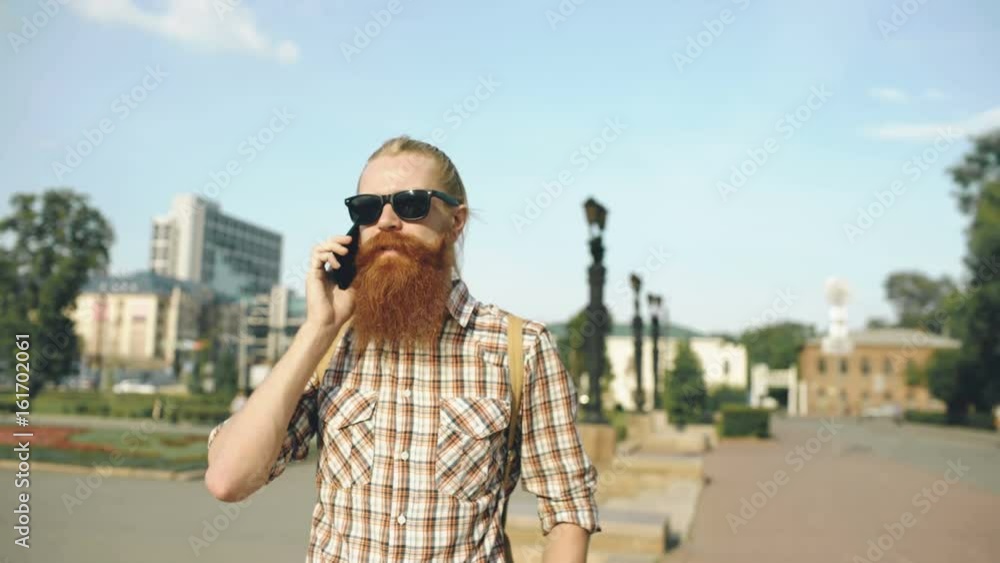 Steadicam shot of Bearded smiling man with backpack walking street and talking phone outdoors Steadicam shot of Bearded smiling man with backpack walking street and talking phone outdoors