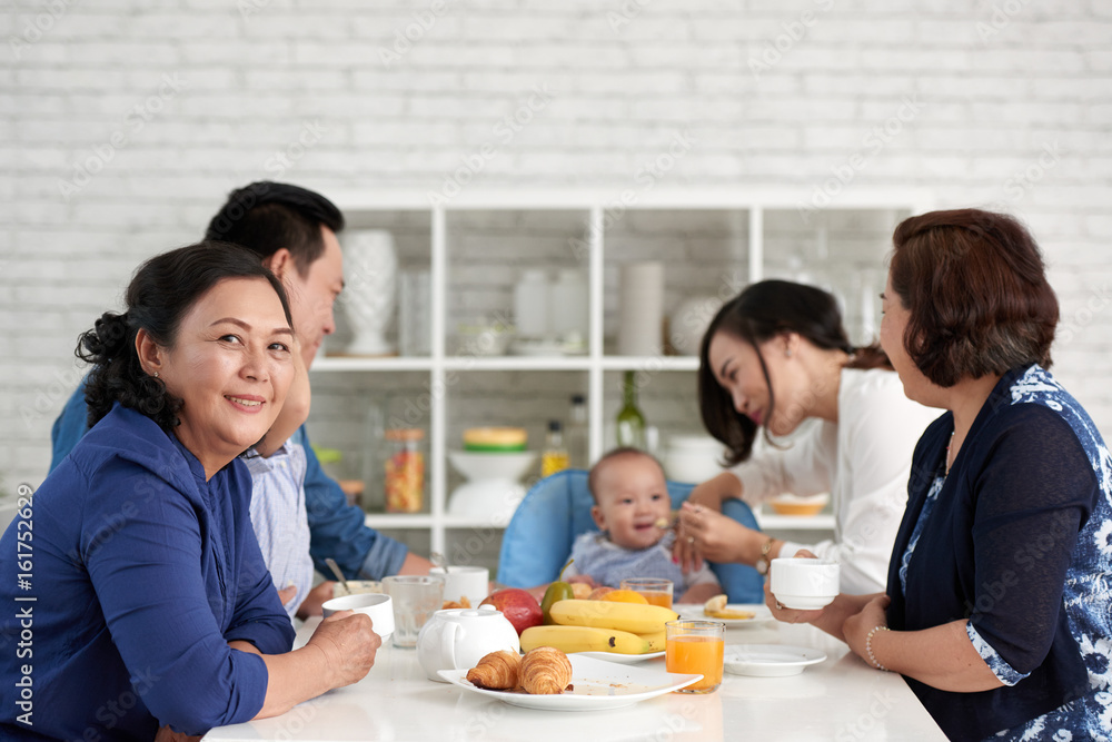 Portrait of mature Asian woman sitting at dining table with big family and smiling to camera Portrait of mature Asian woman sitting at dining table with big family and smiling to camera