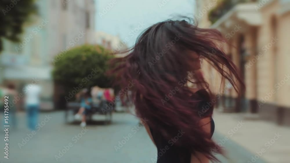 Back view of attractive long-haired curly brunette woman walking down the city-street or alley, turns to camera and gives a beautiful smile, wind plays with her hair. Urban settings on the background. Back view of attractive long-haired curly brunette woman walking down the city-street or alley, turns to camera and gives a beautiful smile, wind plays with her hair. Urban settings on the background.