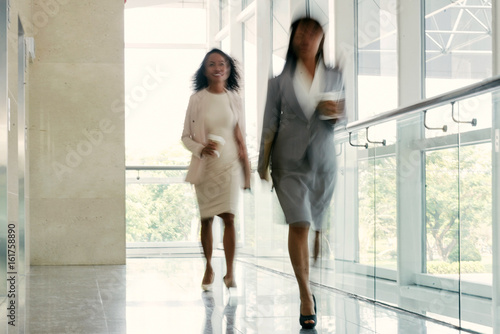Cheerful businesswomen with paper cups of coffee in hands walking down office corridor with panoramic windows, shot in motion