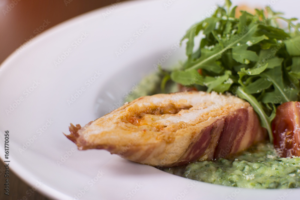 Roasted chicken breast wrapped in bacon slices with greens, tomatoes, and sauce. Beautifully served meal at the restaurant.