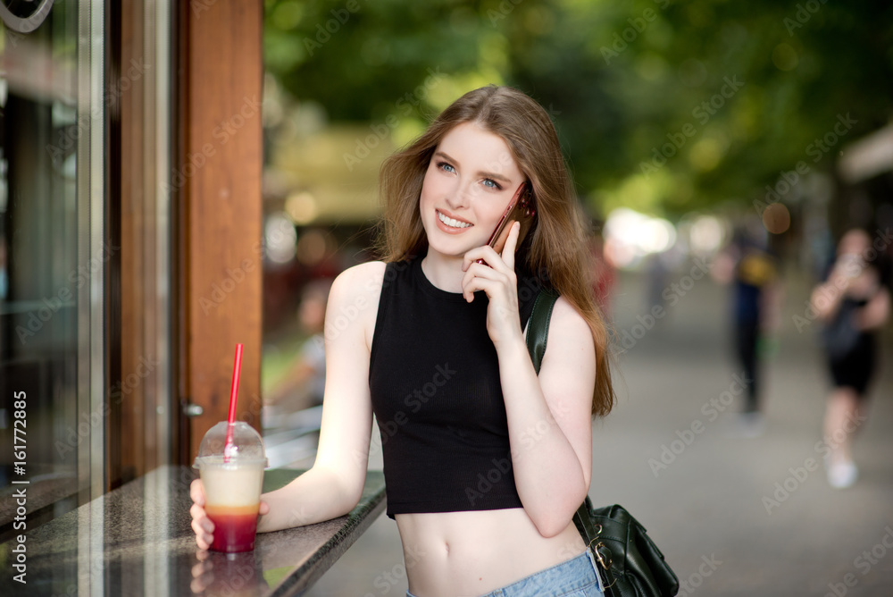Fototapeta premium Young beautiful stylishly dressed girl is walking along the streets of the city, drinking juice, coffee, talking on mobile phone