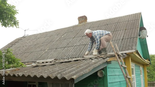 An elderly man is fixing the roof himself. Old wooden house in slate photo