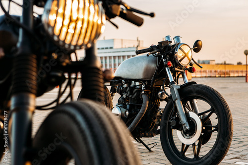 Vintage custom motorcycle cafe racer motorbike with lamp lights turned on. One with grill headlight another with tape cross over optic on empty rooftop parking lot during sunset. Hipster lifestyle.