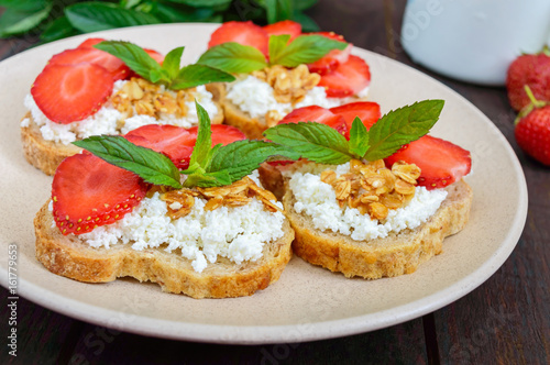 Mini sandwiches with cottage cheese, fresh strawberries, decorated with mint leaves on rye bread  on a dark wooden background. Proper nutrition. Healthy food. Dietary menu