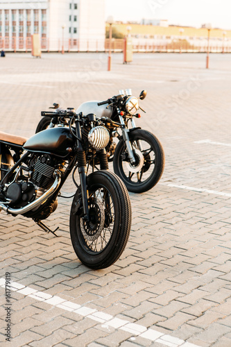 Custom vintage motorbike cafe racer motorcycle with lamp lights turned on. One with grill headlight another with tape cross over optic on empty rooftop parking lot during sunset. Urban lifestyle.